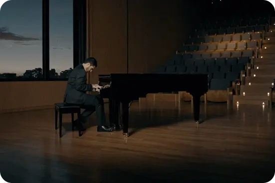 Auditório vazio de madeira com piano de cauda no palco, iluminado por um foco de luz, onde uma pessoa toca.